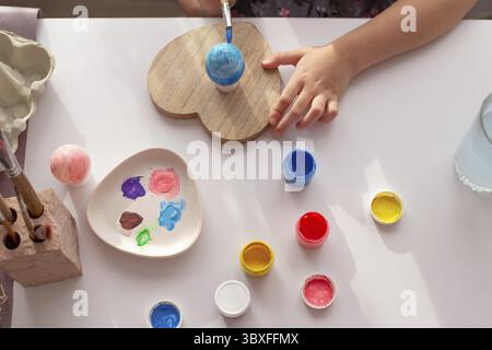 Le mani dei bambini dipingono un uovo su un tavolo bianco con vernici multicolore in vasetti. Vista dall'alto Foto Stock