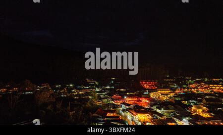 Una splendida vista aerea di un villaggio sul lago di notte, illuminato da calde luci dorate. Lanterne e barche galleggianti creano un modello ipnotico Foto Stock