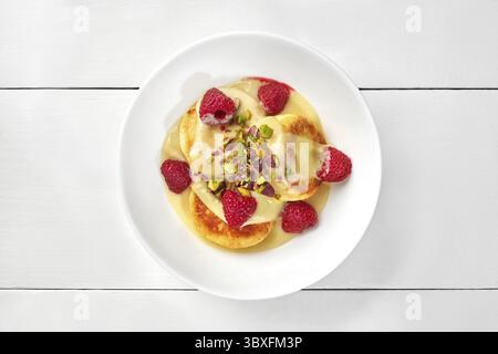 Deliziosi pancake fritti al formaggio dolce cotto serviti con crema di cocco guarnita con lamponi freschi e pistacchi sul piatto, vista dall'alto su wh Foto Stock