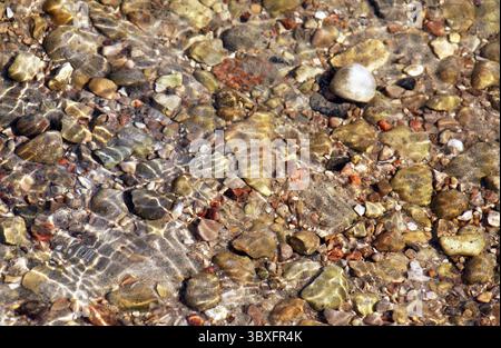 Primo piano di rocce e pietre sotto acque limpide e poco profonde del lago, con la luce del sole che si riflette sulla superficie ondulata. Pietre e ciottoli di diverse dimensioni Foto Stock