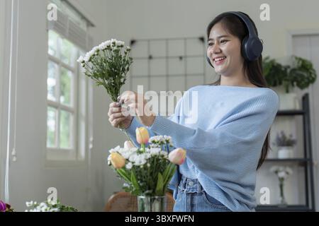 Una giovane donna allegra che indossa le cuffie organizza un bouquet di fiori in una sala luminosa e moderna con luce naturale Foto Stock
