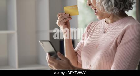 Una donna anziana che esprime emozioni mentre fa shopping online, tenendo una carta di credito gialla in una mano e uno smartphone nell'altra in una casa luminosa Foto Stock