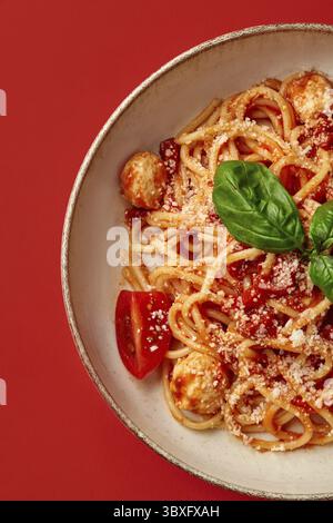 Primo piano di spaghetti con salsa di pomodoro, polpette, basilico e parmigiano, serviti su un piatto su un vivace sfondo rosso Foto Stock