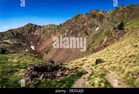 Un paesaggio montano panoramico caratterizzato da aspre cime e un primo piano erboso. Un sentiero roccioso conduce attraverso il verde rigoglioso, con una struttura in pietra in t Foto Stock