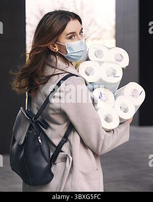 La giovane donna che indossa la maschera di protezione e gli occhiali da sole contro il coronavirus COVID-19 contiene molti fasci di rotoli di carta igienica per la strada della città. Conc Foto Stock