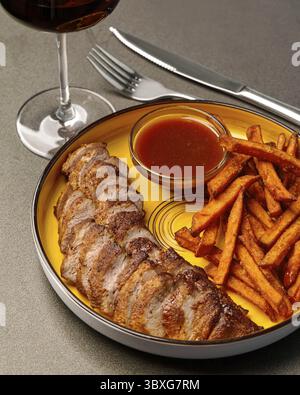 Bistecca di maiale grigliata servita su un piatto giallo con patatine fritte croccanti e un contorno di piccante salsa barbecue all'ananas, accompagnata da un bicchiere di vino rosso Foto Stock