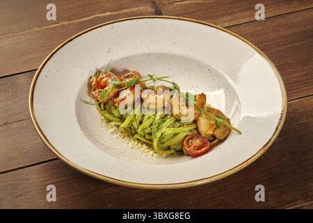 Pasta verde con gamberetti e pesto, servita su un piatto bianco su un tavolo di legno, guarnita con pomodori ciliegini e parmigiano grattugiato Foto Stock