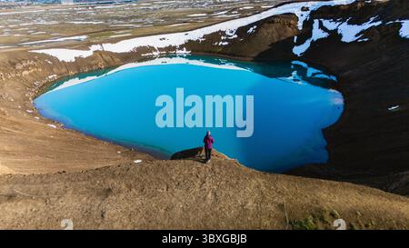 Il maestoso cratere di Krafla presenta una vista mozzafiato con le sue acque blu profonde adagiate su un terreno vulcanico spettacolare. Una figura solitaria ammira la bellezza naturale che caratterizza il paesaggio unico delle Icelands. Foto Stock
