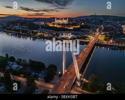 Vista aerea del ponte SNP illuminato che si estende attraverso il tranquillo fiume Danubio verso l'iconico castello di Bratislava sotto il cielo crepuscolo, Bratislava, regione di Bratislava, Slovacchia. Foto Stock