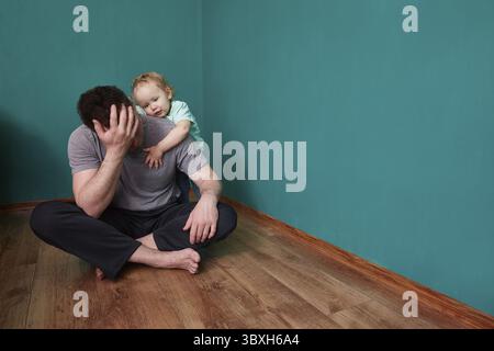 Il bambino si sente dispiaciuto per il padre turbato seduto sul pavimento in una cupa stanza verde Foto Stock