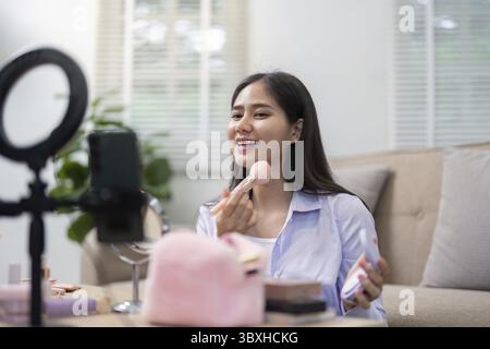 Donna asiatica sorridente che applica il blush con un pennello per il trucco in un ambiente domestico confortevole, usando il suo telefono come specchio Foto Stock