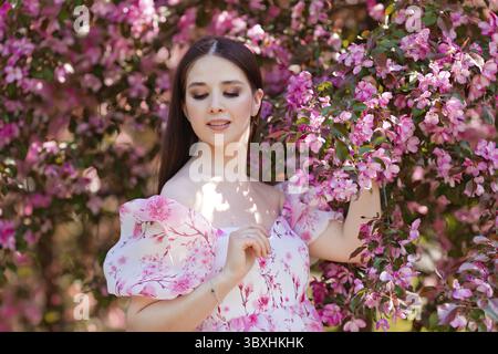 Una bruna in un abito rosa in piedi vicino a alberi di mele in fiore rosa, tenendo un ramo in fiore, in primavera nel giardino, guarda verso il basso. Copia sp Foto Stock