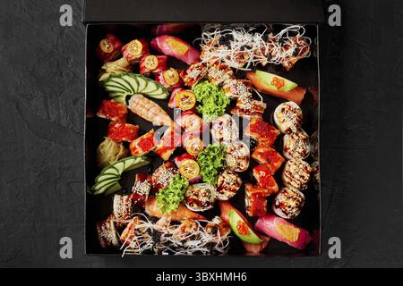 Vista dall'alto di una grande serie di vari panini di sushi giapponesi con zenzero sottaceto, cetriolo fresco, fetta di lime e verdure in scatola di cartone su sfondo nero. Foto Stock