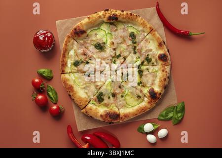 Gustosa pizza con zucchine, prosciutto e mozzarella distesa su carta da forno, circondata da pomodori ciliegini, basilico, peperoncino e palline di mozzarella Foto Stock