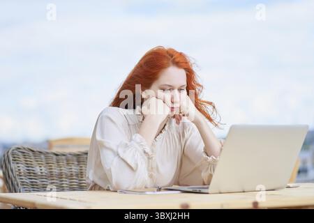 Donna annoiata seduto in un bar sulla strada e che lavora a distanza su un computer portatile. Redhaired uomo d'affari freelance che scrive sul computer Foto Stock