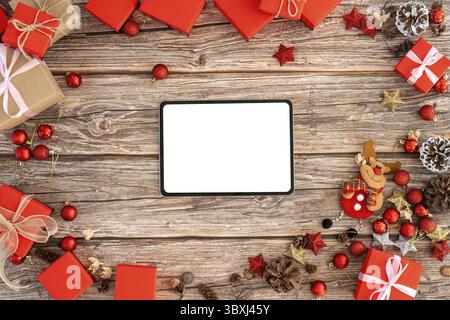 Sfondo natalizio con schermo vuoto per tablet circondato da scatole regalo rosse e decorazioni natalizie su una superficie in legno Foto Stock