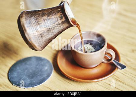 Caffè caldo con vapore in stile turco orientale servito in cezve, una piccola pentola con manico lungo con labbro e tazza su un tavolo di legno Foto Stock