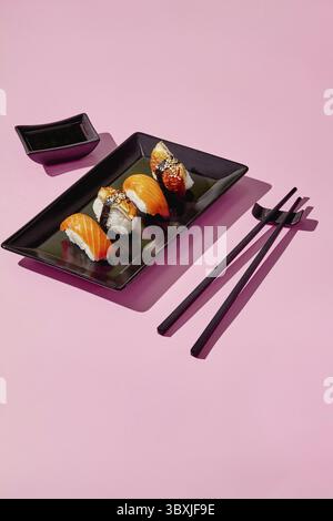 Sushi fresco servito su un elegante piatto nero, accompagnato da salsa di soia e bacchette, creando una vivace scena culinaria sullo sfondo rosa Foto Stock