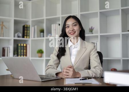Avvocato donna sicura di sé in un ufficio professionale, seduta a una scrivania con un computer portatile e scaffali sullo sfondo Foto Stock