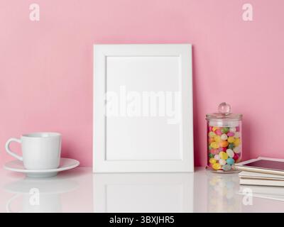 Vuoto telaio bianco e candys in vaso, una tazza di caffè o di tè su un tavolo bianco contro la parete rosa con spazio di copia Foto Stock