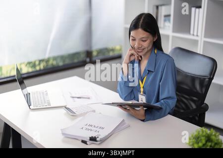 Donna d'affari che esamina i documenti alla propria scrivania con un notebook in un ufficio moderno, sottolineando l'analisi professionale e la produttività Foto Stock