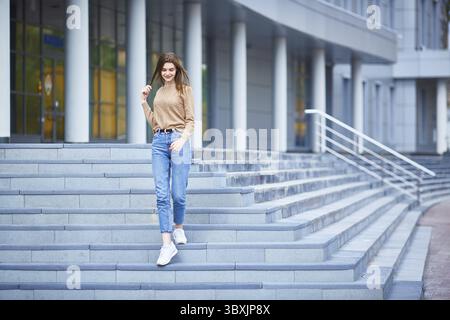 Una giovane donna cammina lungo i gradini di un edificio grigio in una fredda giornata estiva o in un caldo autunno Foto Stock