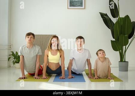 Una bella donna snella, un ragazzo e due adolescenti eseguono esercizi di yoga mentre si sdraiano su un tappetino in posa cobra in uno studio soleggiato. copia spazio Foto Stock