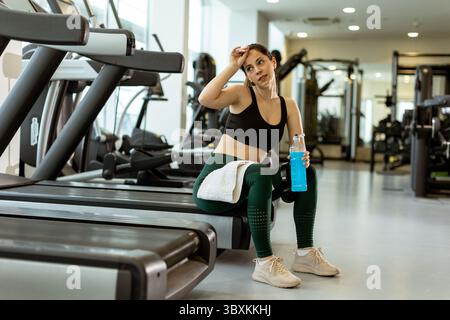 Una donna si prende una pausa da un intenso allenamento cardio in una palestra ben attrezzata, idratando e catturando il respiro mentre è seduta su un tapis roulant. Foto Stock
