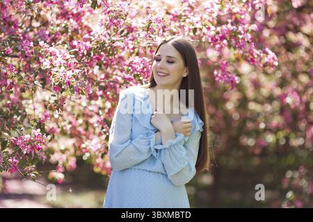Una ragazza felice sorridente, in abito azzurro, con lunghi capelli scuri, in piedi in un giardino rosa fiorito. Primo piano Foto Stock