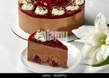 Fetta di cheesecake al cioccolato con spugna umida e ciliegie dolci ricoperte di gelatina di frutti di bosco, panna montata e scaglie di cioccolato, sullo sfondo Foto Stock