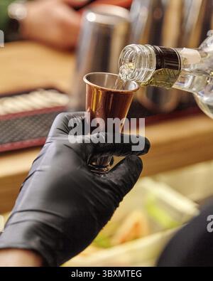 Barman mano in guanto nero versare sapientemente bevande alcoliche chiare dalla bottiglia in scintillante jigger di rame, misurando gli alcolici per garantire un ba perfetto Foto Stock