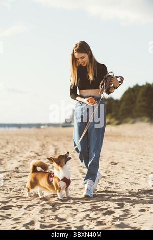 Bella ragazza che gioca con il cucciolo di Corgi che cammina lungo la spiaggia dell'oceano. Donna che guarda al cane il giorno del sole sulla costa oceanica. Stile di vita attivo, animale domestico Foto Stock