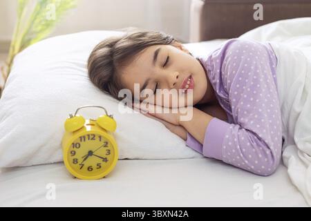 Una bruna che dorme sorridente in pigiama a pois viola giace su un cuscino bianco, su un letto, alla luce del mattino, una sveglia gialla si trova nelle vicinanze Foto Stock