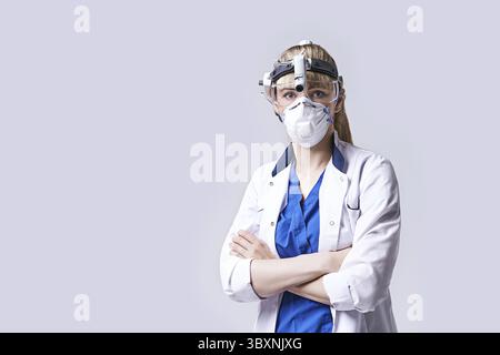 Medico ENT sicuro di sé che indossa la lanterna testa del faro chirurgico, maschera protettiva per il viso e occhiali. Ritratto di una femmina otolaringologa in piedi con braccia cros Foto Stock