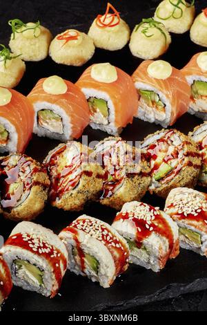 Speciali sushi roll disposti su un tavolo da servizio in ardesia nera. Vista dettagliata degli uramaki ricoperti di salmone e anguilla, tempura fritta croccante e cotta al forno Foto Stock