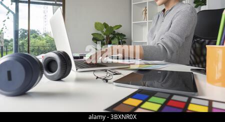 Un grafico che lavora su un notebook, circondato da tavolozze di colori e strumenti di progettazione in un ambiente moderno Foto Stock
