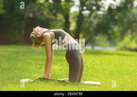 Una donna snella con un top grigio e leggings, in piedi su un tappetino sportivo in estate, sull'erba verde del parco e facendo yoga in posa di cammello. C Foto Stock