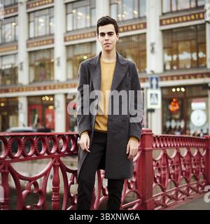 Bell'uomo alla moda, bruna in elegante cappotto grigio, si trova sulla strada nel centro storico di San Pietroburgo. Giovane uomo con capelli scuri Foto Stock