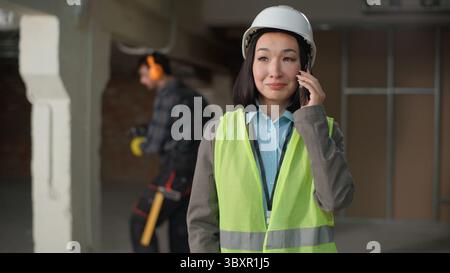 Costruttori multietnici donna coreana uomo caucasico che lavora ingegneri civili in elmetto uniforme caposquadra ispettore che parla telefono cellulare Foto Stock