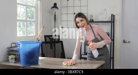 Una donna che indossa un grembiule pulisce una scrivania con un flacone spray e un panno in un ambiente luminoso e moderno Foto Stock