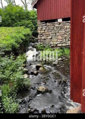 Fagervik, Finlandia: Ruscello che scorre oltre un fondamento in pietra sotto un fienile di legno rosso, circondato da una fitta vegetazione estiva. Foto Stock