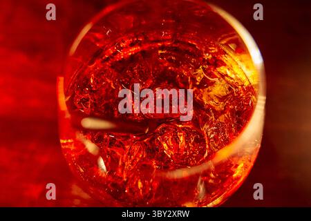 Primo piano di un cocktail color ambra con cubetti di ghiaccio che brillano in un bicchiere, su uno sfondo rosso intenso, creando un'atmosfera calda e invitante. Foto Stock