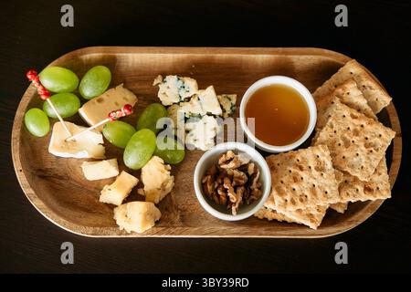 Un vassoio di formaggi gourmet con un assortimento di formaggi, uva verde, cracker croccanti, miele e noci, elegantemente disposte su un vassoio di legno Foto Stock