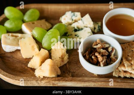 Un piatto gourmet con formaggi assortiti, uva verde, noci, miele e cracker, splendidamente disposto su un vassoio di legno, ideale per l'intrattenimento Foto Stock