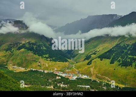 Valle panoramica di montagna con basse nuvole e un paesaggio verde che circonda un piccolo paese situato in una profonda valle alpina, Foto Stock