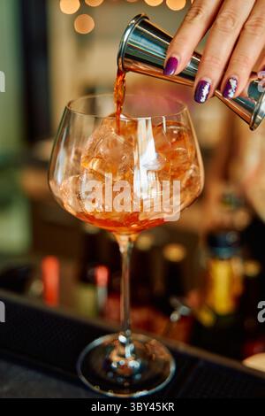 Un primo piano di una mano con unghie in stile che versano un aperitivo su cubetti di ghiaccio in un grande bicchiere di vino, creando una bevanda rinfrescante in un bar moderno Foto Stock