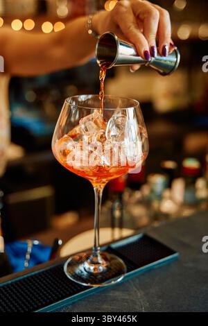 Un barista versa sapientemente aperol in un bicchiere riempito di cubetti di ghiaccio su un bancone, creando un rinfrescante cocktail aperol spritz con precisio Foto Stock