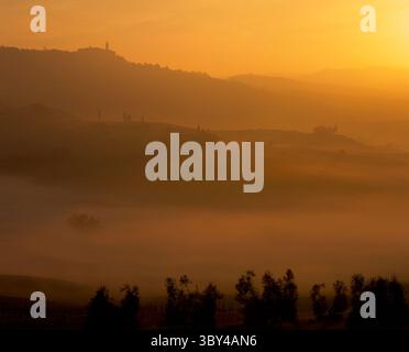 La nebbia percorre le valli all'alba, mentre la calda luce dell'alba prende la cattedrale in cima alla collina di Pienza con cipressi nel fi Foto Stock