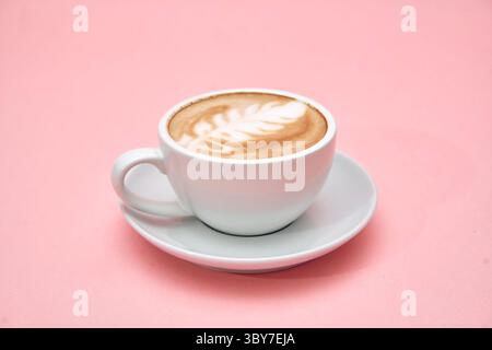 Un latte sereno con un'intricata schiuma art si trova in una tazza e un piattino in ceramica bianca su un morbido sfondo rosa, che esemplifica l'eleganza e il caffè gourmet Foto Stock