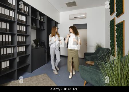 Due donne si impegnano in una vivace conversazione sulle iniziative per la salute delle donne in un ufficio elegante pieno di verde e file organizzati, mostrando un Foto Stock